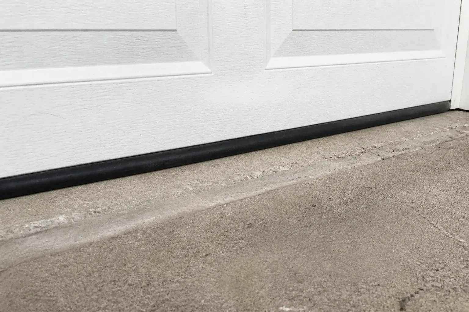 Black bottom seal along a closed white garage door at a concrete threshold. — Garage Door Bottom Seal Replacement, Georgina