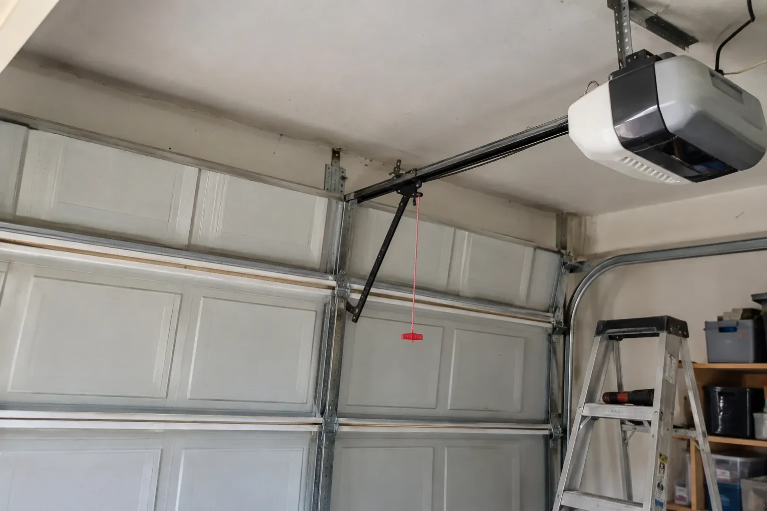 Garage-door opener on the ceiling with a bent black arm to a paneled door, a red release cord and a stepladder.